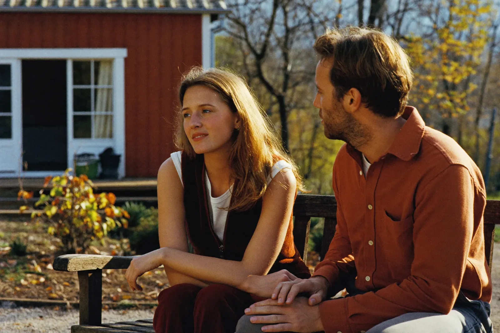 A couple on a garden bench in autumn light - he is turned toward her, she gazes into the distance