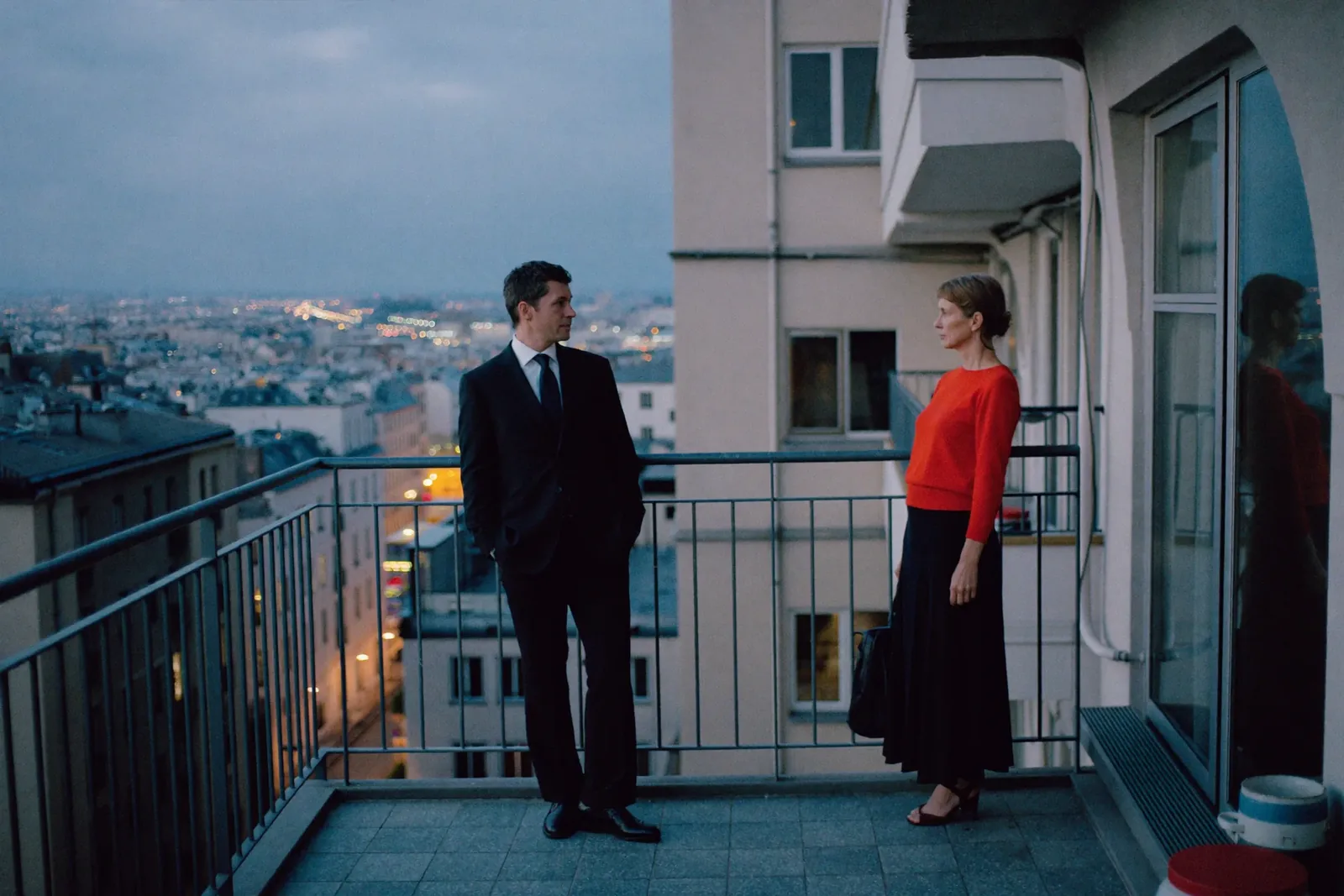A couple standing apart on a balcony at dusk, one turned toward the other