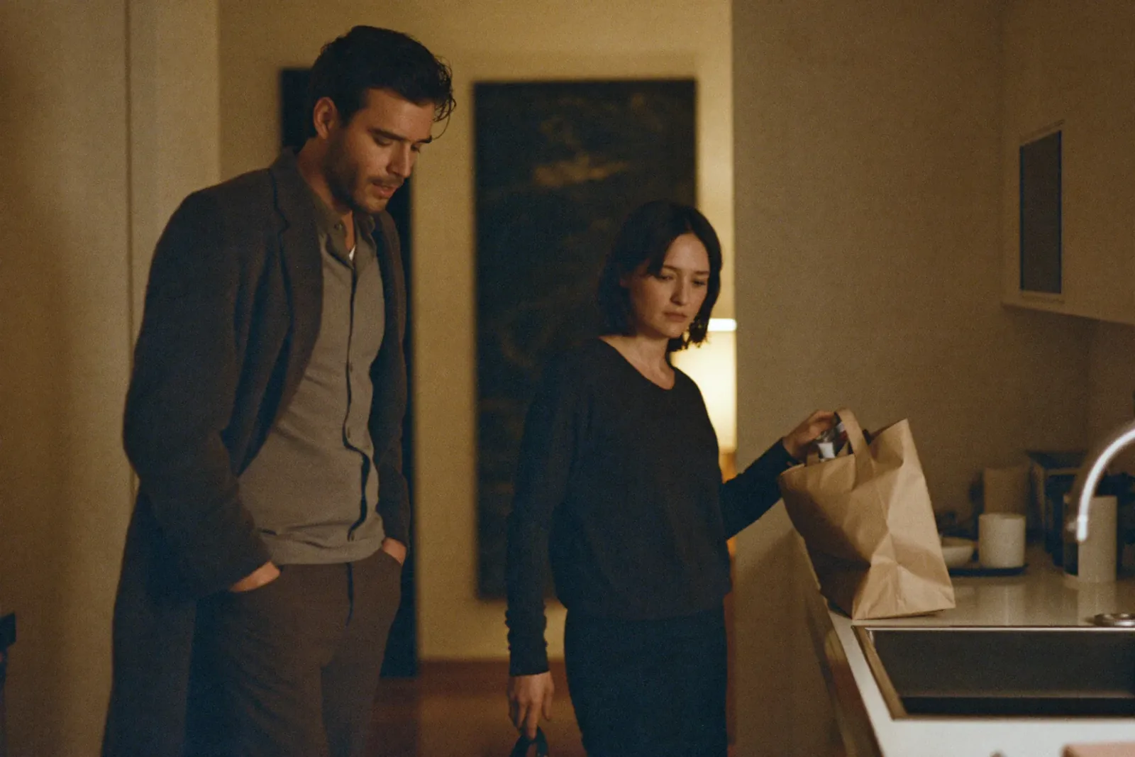 An evening kitchen: he stands with hands in pockets, she unpacks a paper bag, both looking down