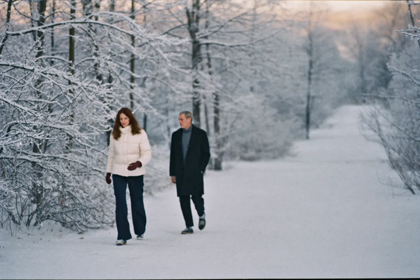 A couple walking a snowy path a few steps apart, in winter light