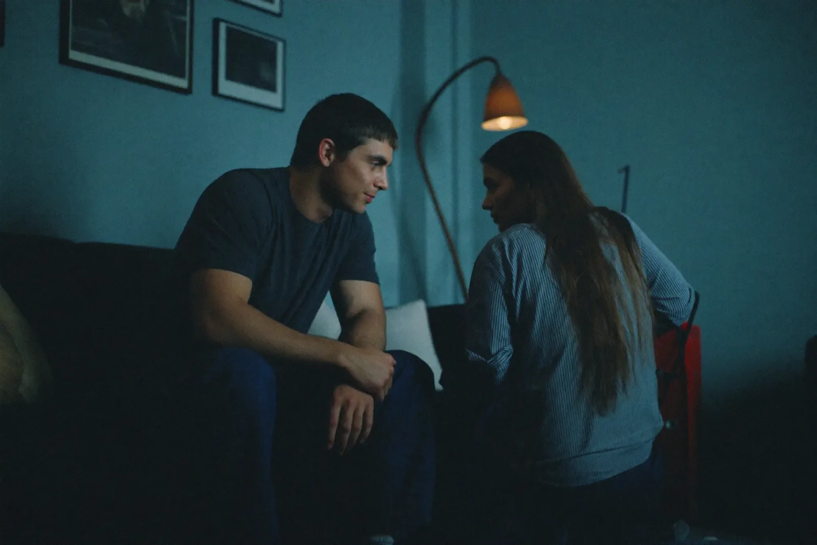 A dim blue evening room: she sits turned away, he watches her from the couch