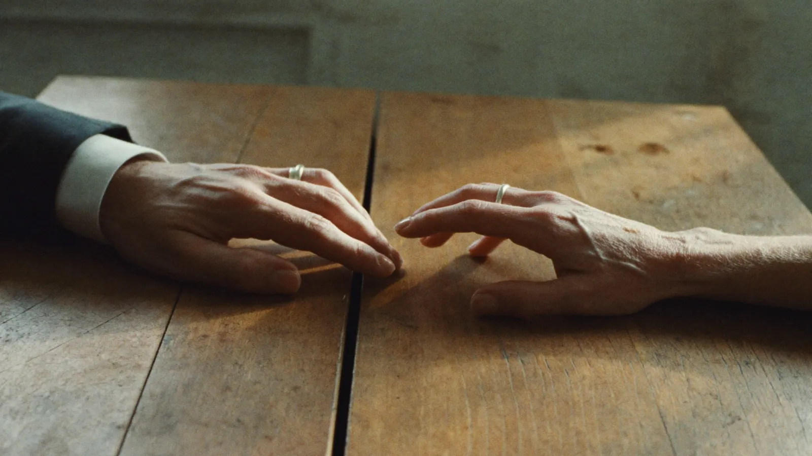 Two hands with wedding rings resting on a table, fingers a breath apart